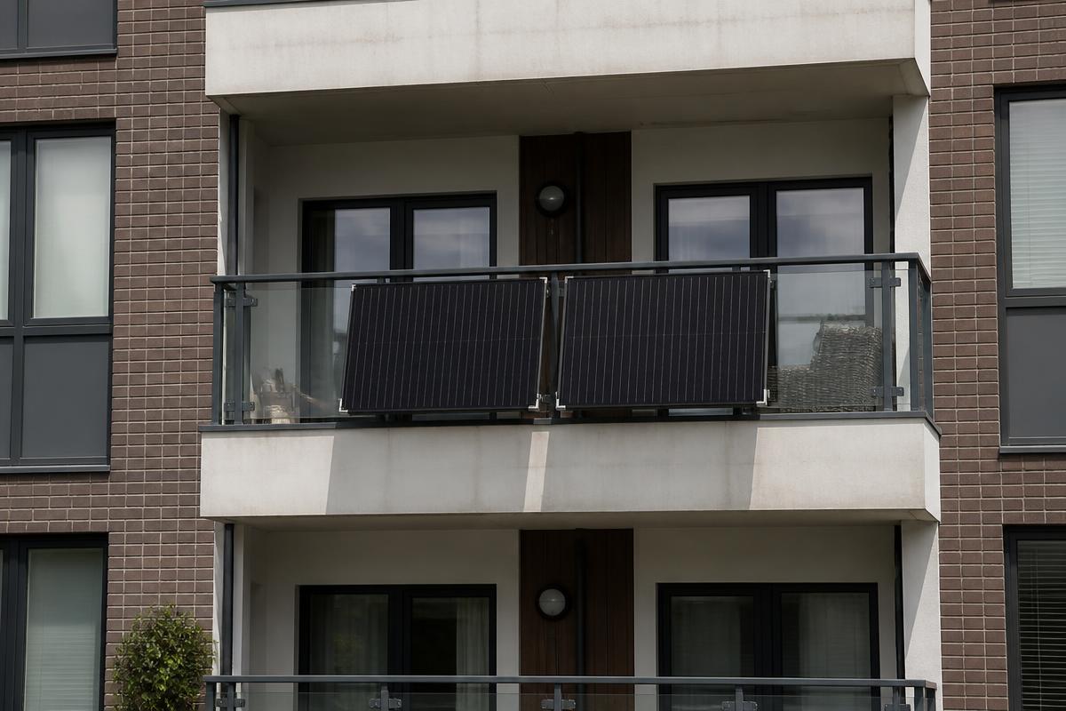 Two slim black solar panels bracketed to the outside of an apartment balcony railing, seen from outside the building